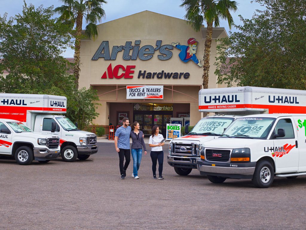 A U-Haul dealer escorts a Caucasian couple to the parking lot of an ACE Hardware filled with U-Haul trucks and vans.