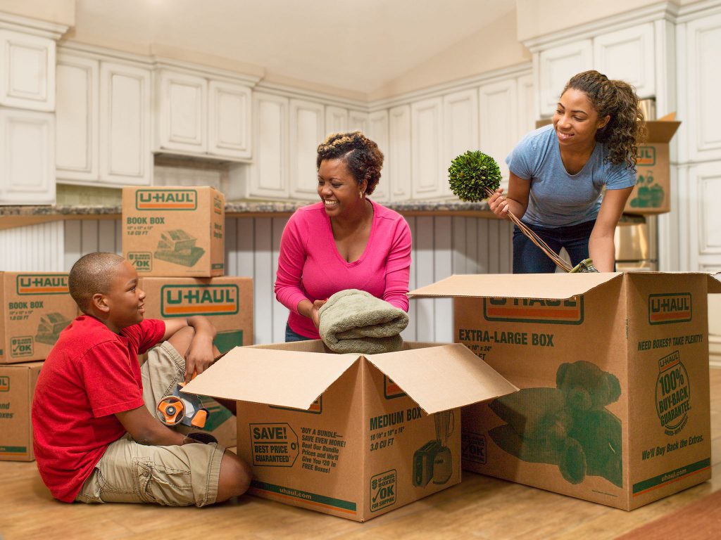 A family packing and moving their things. A mother, daughter and son are seen putting a blanket and furniture piece into medium and extra-large moving boxes.