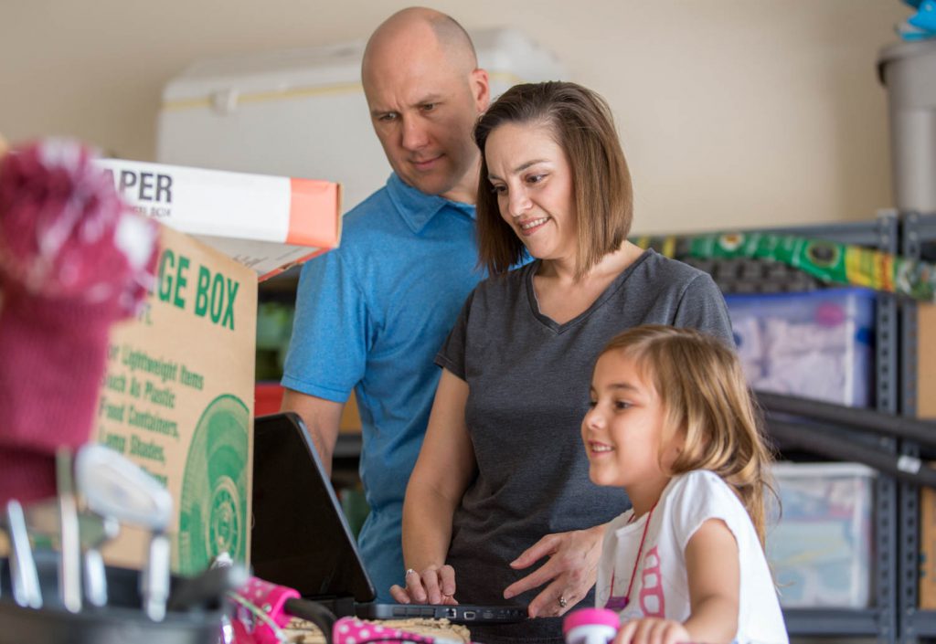 A family looking at a laptop and they are in the process of moving. The dad, mom and daughter are all smiling with their belongings surrounding them. They are looking into Truck Share 24/7 a self-service moving experience provided by U-Haul.