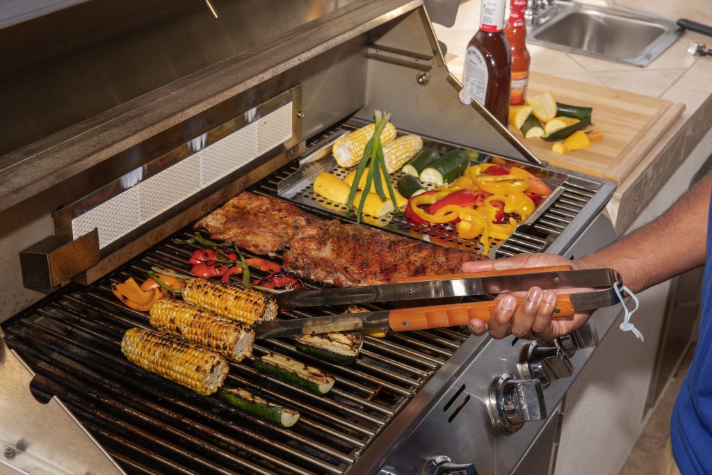 A propane-powered grill being used to cook corn, bell peppers, zucchini, meat, and squash. BBQ and buffalo sauces wait nearby to be dripped, smothered, and enjoyed with the freshly grilled food on game day.