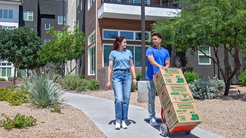 A Moving Help provider pushes a dolly carrying U-Haul moving boxes while talking to the customer outside of her apartment 