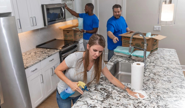 A woman cleans her apartment as two moving labor providers pack her kitchen items into U-Haul moving boxes