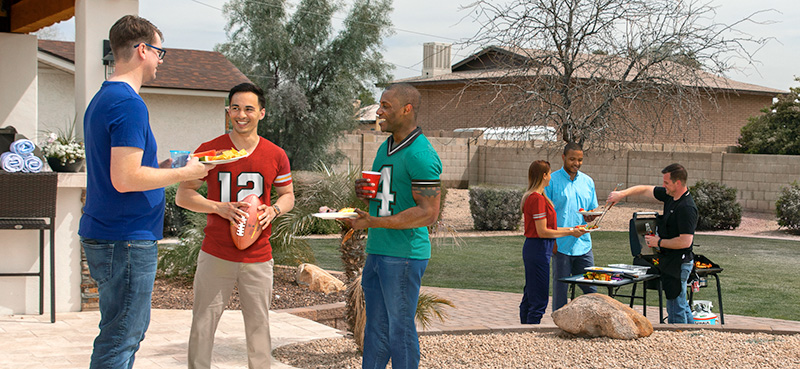 The setting is a large, backyard hosting for NFL opening day bbq. In front, 3 men in football jerseys are enjoying a chat with plates of food. One man in the background is grilling with U-Haul propane and serving up the food on plates.