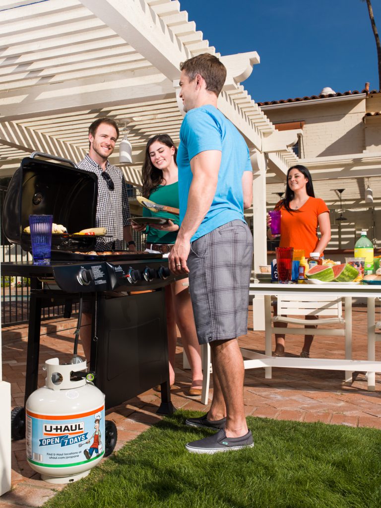 Couples bbq in backyard with the U-Haul propane tank on the side of the grill. A lady is at the picnic table with food and watermelon atop, and friends talking around the grill with drinks